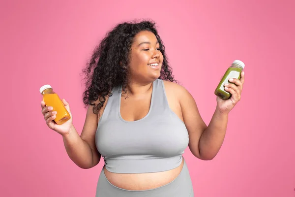 stock image Happy african american oversize woman in sportswear holding jars of juice or smoothies isolated on pink background, studio shot. Body care, vitamins for sports