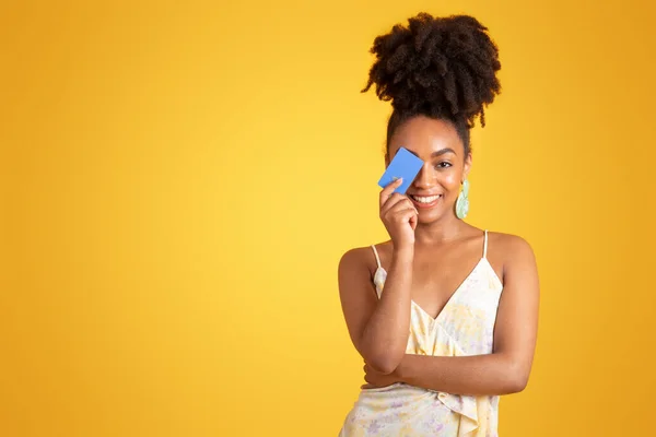 stock image Happy young african american woman in dress covering eye with credit card, isolated on yellow studio background. Shopaholic and sale, finance, money, lifestyle, ad and offer