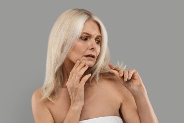 Kuru Sonu. Stresli, olgun bir kadın, sakat saçlarına bakıyor. Gri stüdyo arka planına sarılıp havluya sarmalanarak güzellik sorunları yaşayan endişeli yaşlı bir bayan.