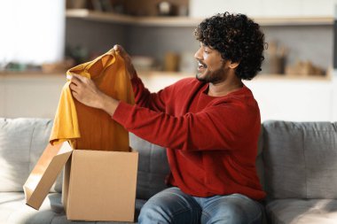 Başarılı Çevrimiçi Alışveriş. Doğulu neşeli bir erkek müşteri elinde yeni kıyafetler, moda mağazasından kutuları açıyor ve evdeki koltukta otururken sarı tişörtünü çıkarıyor.