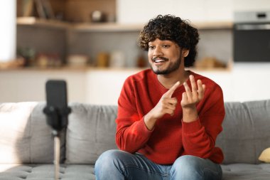Blog Kavramı. Yakışıklı Hintli adam evde akıllı telefonuyla video kaydediyor, gülümseyen genç doğu erkeği sosyal ağlar için içerik yakalıyor, telefonda konuşuyor, yer kopyalıyor.