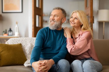Gülümseyen Avrupalı olgun bir çift, yaşlılıkta aile rahatlığı ve aşk ilişkisinin tadını çıkarıyor, koltukta oturuyor, evine bakıyor. Eşler Oturma Odasında Birlikte Rahatlıyor