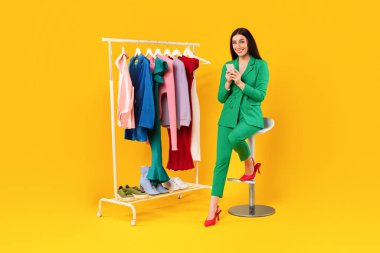 İnternet üzerinden iletişim kurulamıyor. İnternetten alışveriş yapmak için cep telefonu kullanan mutlu Avrupalı kadın, elbise rafının yanındaki sandalyede oturuyor, sarı stüdyo arka planında yeni parlak kıyafetler seçiyor..