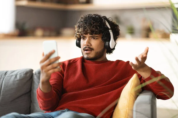 自宅でソファに座っている間にスマートフォンの画面を見てヘッドフォンで欲求不満若いインド人男性 携帯電話で悪いメッセージを読んで不安東部の男 感情的にニュースに反応 — ストック写真