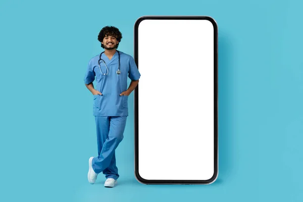 医务工作者兴高采烈 英俊的卷曲的印度年轻医生 拿着大手机摆姿势 白色的空白屏幕 蓝色背景隔离 医生推荐远程保健应用程序 复制空间 — 图库照片