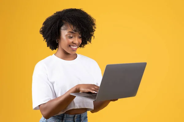 オレンジ色のスタジオの背景に隔離されたラップトップ上のカジュアルなタイピングでかなり千年の黒の女性を笑顔 仕事や研究のためのデバイスリモートで ブログや通信 ソーシャルネットワーク — ストック写真
