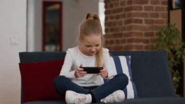 Modern çocukluk eğlencesi. Akıllı telefonda video oyunu oynayan heyecanlı küçük kız, başarısını kazanıyor ve eğleniyor, evde eğleniyor, çekim takibi, yavaş çekim, serbest alan