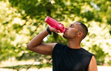 Ciddi sporcu bir zenci yaz parkındaki parkta bir şişe su içiyor. Spor ve susuzluk, açık hava sporları, kardiyo egzersizleri, rekabet ve yaşam tarzı, su dengesi.