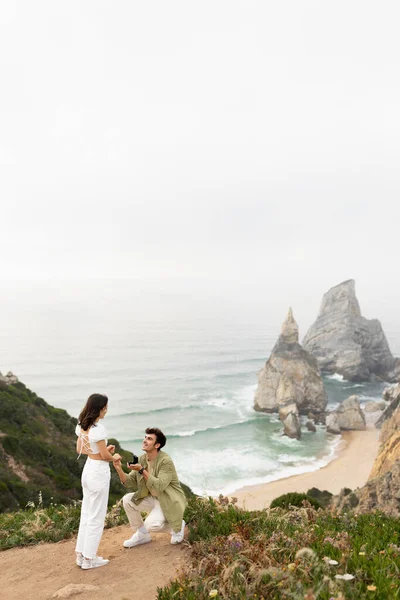 彼女は上にはいと言った 海の岩の崖の上に立っている若いカップル 男は彼の膝の上に女性に提案立って 息をのむような海岸線の景色 垂直ショット フリースペース — ストック写真