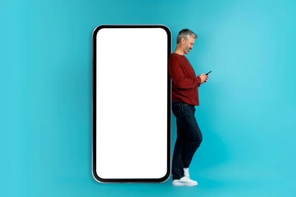 最新的移动应用程序概念 白发苍苍 40多岁的中年男子 穿着休闲装 用手机 站在大手机旁边 — 图库照片