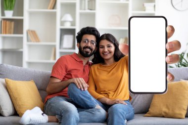 Yeni Mobil Uygulama Reklamı. Elinde boş ekran olan akıllı telefonu tutan mutlu Hintli çift, model için boş alanı olan aygıtı gösteriyor, oturma odasında iç tarafta birlikte oturuyor, kolaj yapıyor.