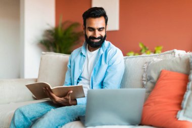 Çevrimiçi Eğitim. Orta Doğulu serbest çalışan bir adam kitap okuyor, dizüstü bilgisayara bakıyor, çalışıyor ve internette çalışıyor. Evdeki koltukta oturuyor. Adam hafta sonları bilgisayarda öğreniyor. E-Öğrenme, Web Kursu