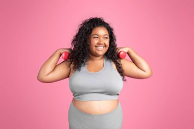 Spor giyim sektöründe pozitif kadın dambıllarla el egzersizi yapıyor. Pembe stüdyo arka planında spordan keyif alıyor. Aktif yaşam tarzı ve vücut bakımına uygun