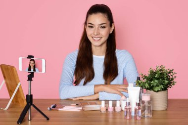 Neşeli, güzel, İspanyol asıllı, günlük kıyafetlerle gezen genç bir kadın güzellik blogcusu, profesyonel blog yazarı seti ve akıllı telefon kullanıyor, telefon kamerasına gülümsüyor, pembe arka plan, kopyalama alanı.