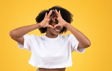 Turuncu stüdyo arka planında izole edilmiş, elleri ile kalp işareti yapan gündelik siyahi kadın. Eğlence, aşk, romantizm insani duygular, yaşam tarzı ve moda, reklam ve teklif