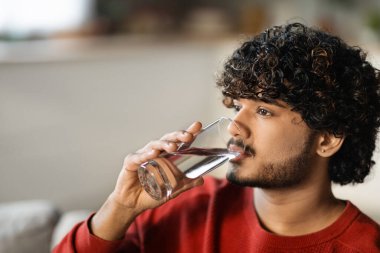 Sağlık Konsepti. Genç Hintli bir erkeğin evde dinlenirken bardaktan su içmesi, yakışıklı bir doğu erkeğinin evde dinlenirken içkisini tazelemesi, boşluğu kopyalaması.
