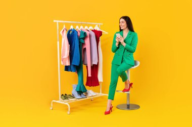 Harika bir alışveriş uygulaması. Cep telefonu kullanan, gardırop rafındaki eşkıya kıyafetlere bakan, sarı arka planda oturan heyecanlı genç bayan.