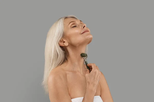 惊吓疗法 用绿石翡翠滚筒做颈部部位的漂亮女人 站在灰暗的背景下 快乐的老年女性用漂亮的工具做举重按摩 — 图库照片