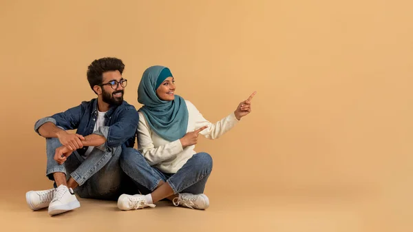 コピースペースを脇に置く幸せなイスラム教徒の家族 広告のための無料の場所を示すヒジャブで陽気なアラビア人の男と女 ベージュの背景に座っているイスラム教徒のカップル パノラマ — ストック写真