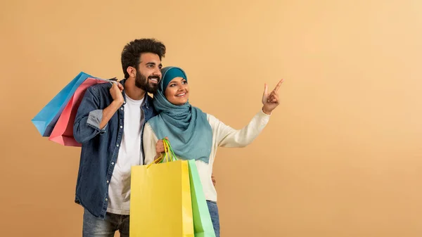 Reklamní Nápis Happy Muslim Pár Držení Nákupních Tašek Ukazující Aside — Stock fotografie