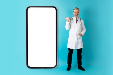 Neşeli, yakışıklı, orta yaşlı adam doktor mavi stüdyo arka planında izole edilmiş beyaz ekranlı akıllı telefonu işaret ediyor. Pratisyen hekim tele sağlık uygulaması, model, tam boy öneriyor.