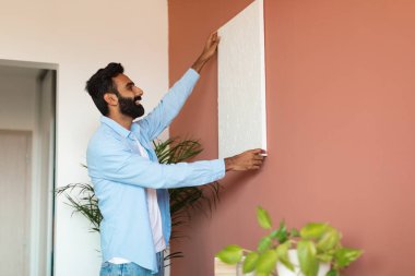 Ev dekorasyonu. Modern ev içinde duvarlara ahşap resim çerçevesi asan gülümseyen Arap adamın yan görüntüsü. Oturma odasında boş poster tutan sıradan bir adam, kopyalama alanı