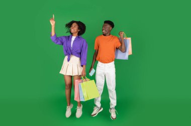 Gülümseyen Afrikalı genç Afro-Amerikalı erkek ve kadın alışveriş kolikleri, alışveriş torbaları, havada donma, boş alanda işaret parmağı, yeşil stüdyo arka planında izole edilmiş. Boş zaman, satış, alışveriş.