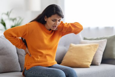 Güzel, genç Hintli kadın acı içinde rahat bir kıyafet giyer evdeki oturma odasındaki koltukta oturur, boynuna ve sırtına dokunur, kas gerilmesinden, skolyozdan ya da kırışıklıklardan muzdarip olur, fotokopi çeker.