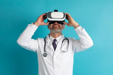 Doktor orta yaşlı, gri saçlı, VR kulaklık takan, mavi stüdyo arka planında izole edilmiş, dokunaklı gözlükler takan adam. Sağlık hizmetleri kavramında geleceğin tıbbı ve modern teknoloji aletleri