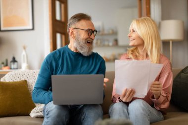 Basit evrak işleri. Evrak tutan, dizüstü bilgisayar kullanan, evdeki rahat oturma odasındaki koltukta oturan mutlu, olgun bir çift. İyi Finans Haberleri Konsepti