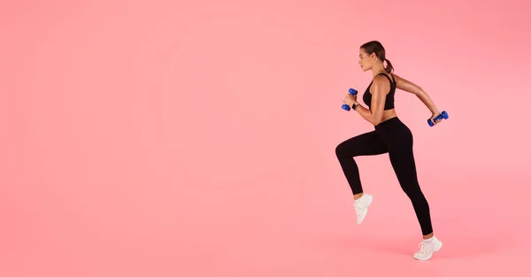 スポーツ動機 手にダンベルを持って走るスポーティ若い女性の動きのショットで ピンクのスタジオの背景に空気中でジャンプスポーツウェアでアスレチック若い女性 コピースペースとパノラマ — ストック写真