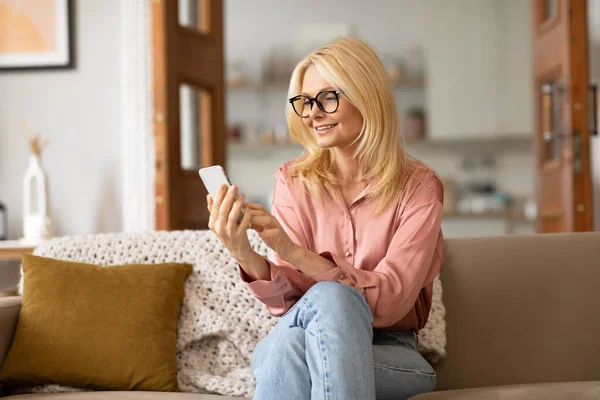移动技术 快乐的老年妇女戴眼镜用智能手机 阅读网上新闻 发短信和通过沙发交流 坐在舒适的客厅里 — 图库照片