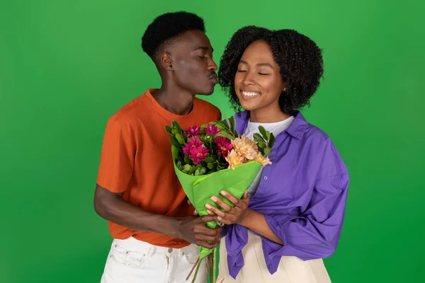 快乐的年轻的非洲裔美国人在脸颊上亲吻女人 送给她一束鲜花 在绿色的工作室背景下被隔离 日期和关系 周年纪念日 节日庆祝活动 — 图库照片