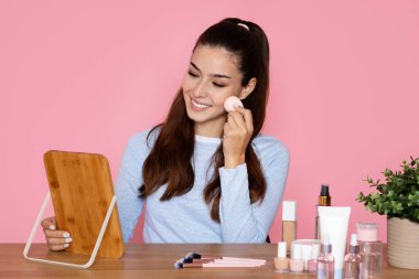 Mutlu neşeli gülümseyen güzel esmer kadın pembe stüdyo arka planında masada oturuyor, aynaya bakıyor, güzellik blogcusu makyaj vakfı uyguluyor ya da allık sürüyor, sünger kullanıyor, boşluğu kopyalıyor.