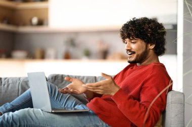 Evde dizüstü bilgisayarla video görüşmesi yapan gülümseyen Hintli genç adam, Millennial East 'in yakışıklı erkeği oturma odasında otururken internet konferansı için bilgisayar kullanıyor, çevrimiçi iletişimin tadını çıkarıyor.