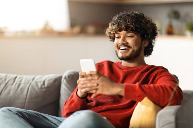 Evdeki koltukta dinlenirken akıllı telefondaki neşeli Hintli Adam Mesajlaşması, Çevrimiçi İletişim ya da Sosyal ağları taramak için cep telefonunu kullanan Mutlu Doğulu Adam, Boşluğu Kopyala
