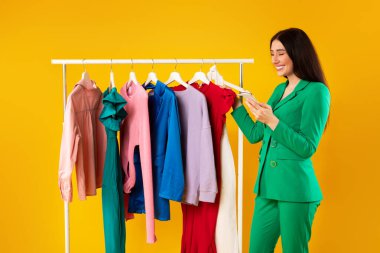 Cep telefonuyla alışveriş yapan mutlu bayanın giyim kuşağının yanında durması, modaya uygun kıyafetler seçmesi ve sarı arka planda stilistle görüşmesi, stüdyo çekimi.