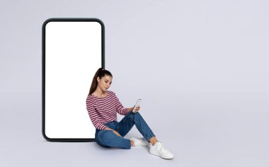 Güzel mobil uygulama. Havalı, neşeli, güzel, esmer, günlük kıyafetler giyen, gri arka planda beyaz ekran, taklit, kopyalama alanı olan, büyük telefon başında oturan uzun saçlı kadın.
