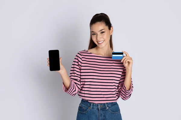 黒の空の画面とグレーの背景に隔離されたプラスチック製の銀行カードで電話を示すカジュアルな服装で肯定的な陽気なかなりブルネットの若い女性 オンラインバンキング ショッピングコンセプト — ストック写真