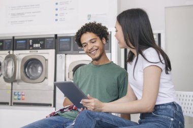 Çamaşır yıkayan ve dijital tablet bilgisayar kullanan mutlu Latin erkek ve kadın çamaşırhane odasının iç tarafında modern çamaşır makinelerinin yanında oturuyorlar. Laundrette Servis Reklamı
