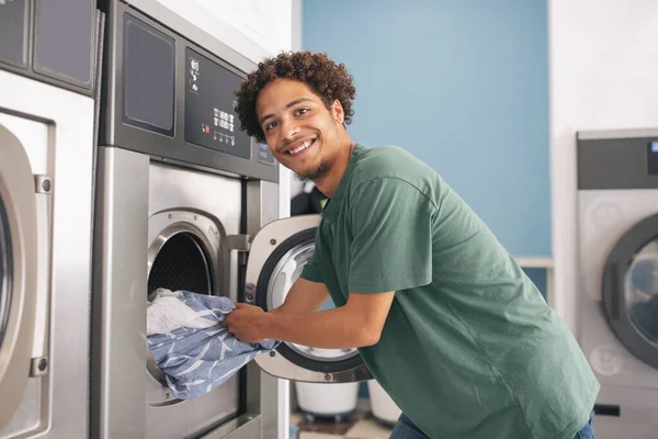 洗衣店服务 笑拉丁小伙子装洗衣机把脏衣服和看相机 贴在洗衣房室内 杂务及方便市民的概念 — 图库照片