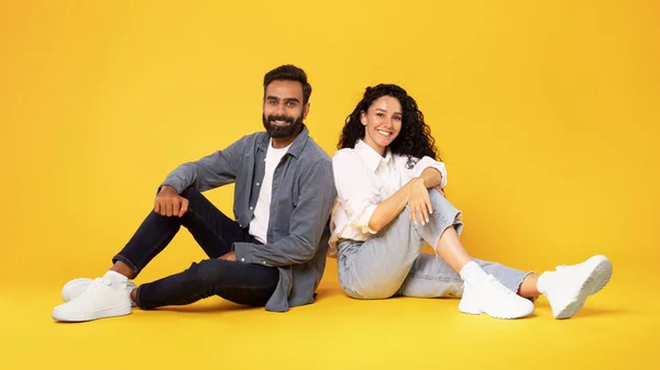 Stock image Romantic Relationship Advert. Cheerful millennial arabic man and woman in white t-shirts sitting on floor together, posing over yellow studio background, panorama. Love and good family offer