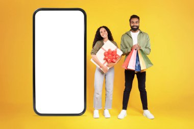 Alışveriş Uygulaması. Sarı Stüdyo Arkaplan 'da boş ekranı olan, hediye kutusu ve renkli alışveriş torbaları olan, büyük cep telefonlu neşeli Arap müşteriler. Mockup, Satış Teklif Reklamı. Kolaj