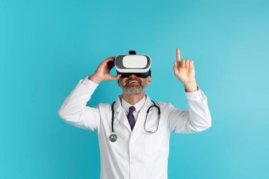 Sanal Gerçeklik Oyunları Sağlık Hizmetleri için. Orta yaşlı hekim doktor sanal gerçeklik kulaklığı kullanıyor, VR gözlüğüne dokunuyor ve üstündeki boşluğu kopyalıyor, mavi arka planda izole edilmiş.