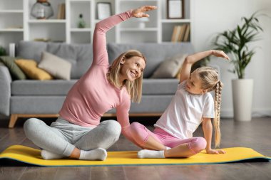Neşeli anne ve kadın çocuk evde birlikte yoga yapıyorlar, anne ve küçük kız oturma odasının iç kısmında spor paspası uzatıyorlar, yan eğilmelerini sağlıyorlar, antrenmanların keyfini çıkarıyorlar.