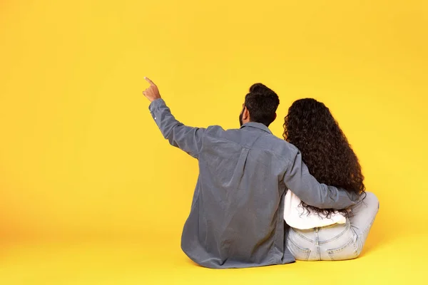 コピースペースで指を指す中東のカップルのバックビュー 黄色のスタジオの背景に座っている広告テキストを抱擁し このオファーのコンセプトをチェック — ストック写真