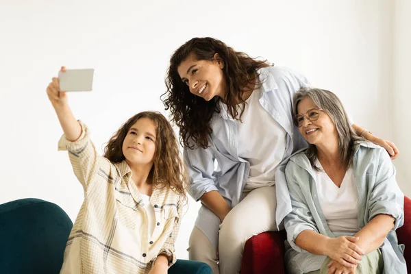 stock image Glad european adult, senior women and teen girl sit on sofa, make selfie on smartphone in living room interior. Family, photo female generation, relationship mom, grandma and daughter, video call