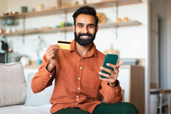 电子商务带来的好处 用电话和信用卡向坐在沙发上在家里拍照的阿拉伯男子微笑购物 网上采购广告银行服务或流动应用 — 图库照片