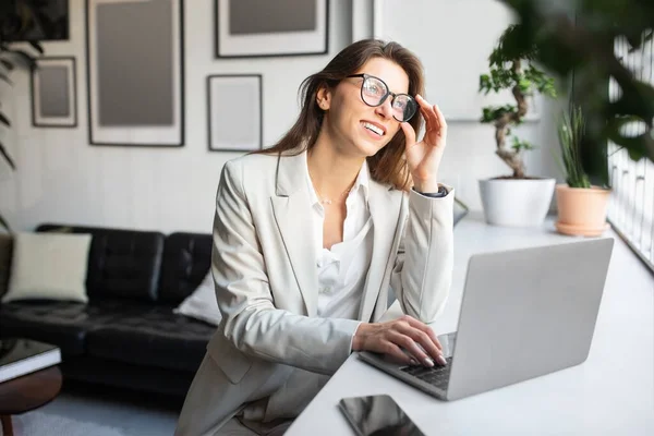 戴着眼镜 带着女企业家微笑 在舒适的工作空间里与笔记本电脑一起工作 凝视着窗户 沉思着 在咖啡店做网上工作的自由职业者 — 图库照片