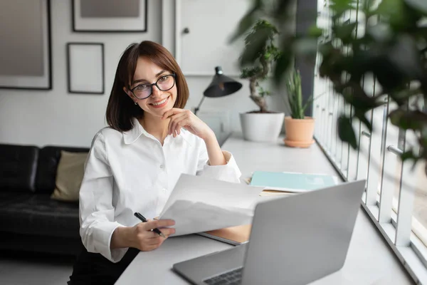 文书工作很简单快乐的女性财务顾问使用笔记本电脑和持有文件 在舒适的办公室内部工作 对着相机微笑 在网上处理文件的女商人 — 图库照片